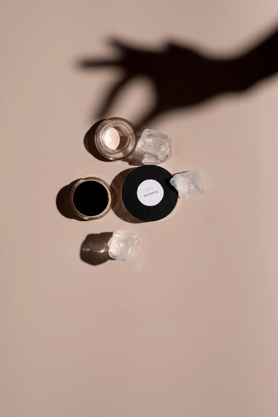 Small glass cosmetic jars and clear crystals on a blush backdrop, with the shadow of a hand reaching in.
