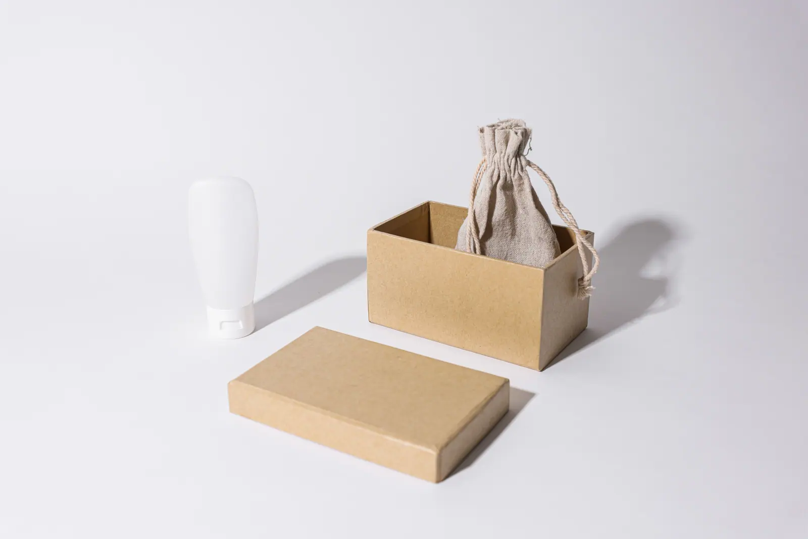 A white squeeze tube, a kraft box and a linen pouch on a pale grey surface.